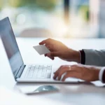 Person holding a card while using a laptop, representing secure online transactions with our cybersecurity solutions.
